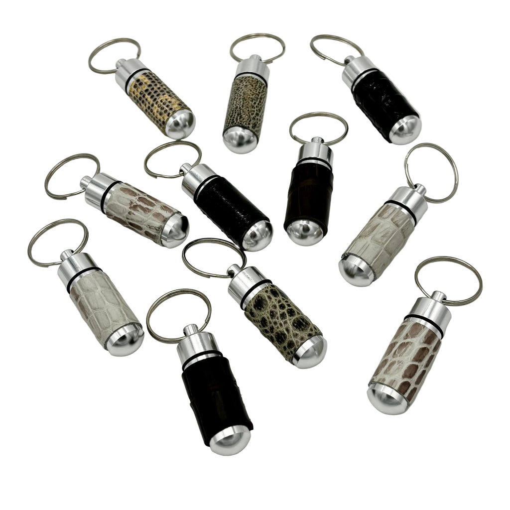 Collection of exotic leather pill capsule keychains in various skins including alligator, python, and black leather with stainless steel hardware